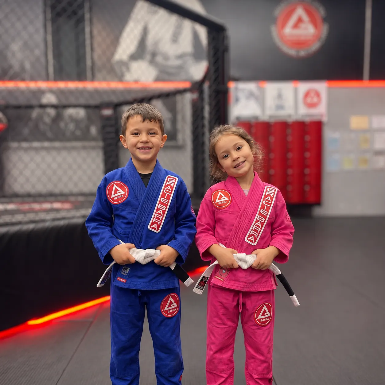 Twee kinderen in een blauw en een roze Gracie Barra-pak staan naast elkaar op de mat bij FPA, met de kooi en de rode lockers op de achtergrond